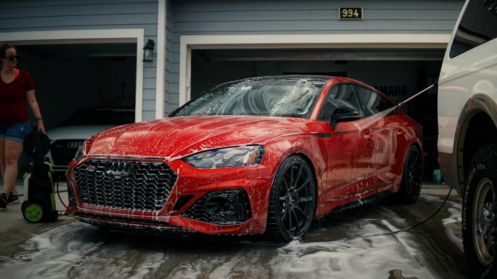 A red sports car is being cleaned with soap in a driveway, featuring a modern garage background.